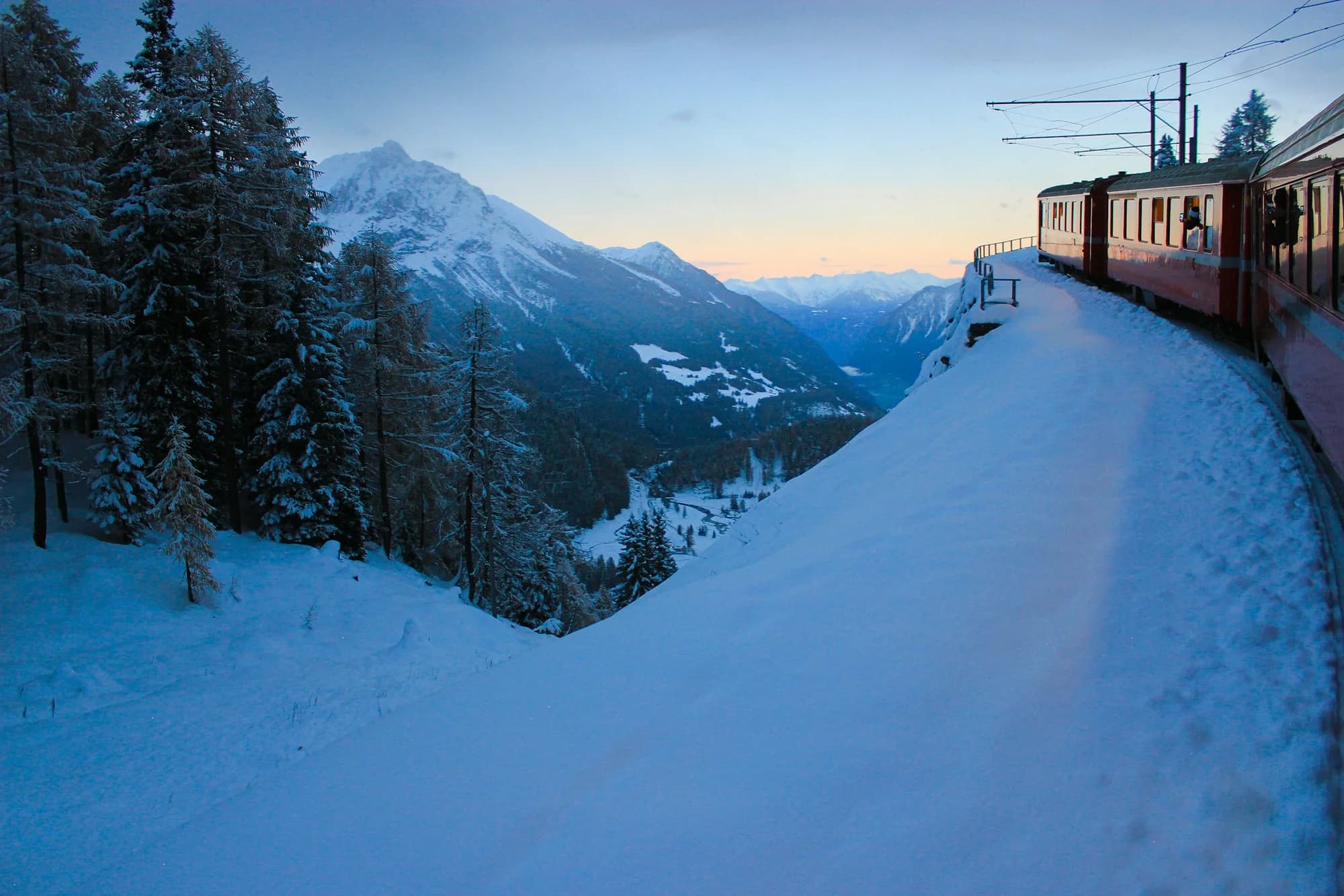 A red Bernina train beside a snow-covered Alpine slope at dusk.
