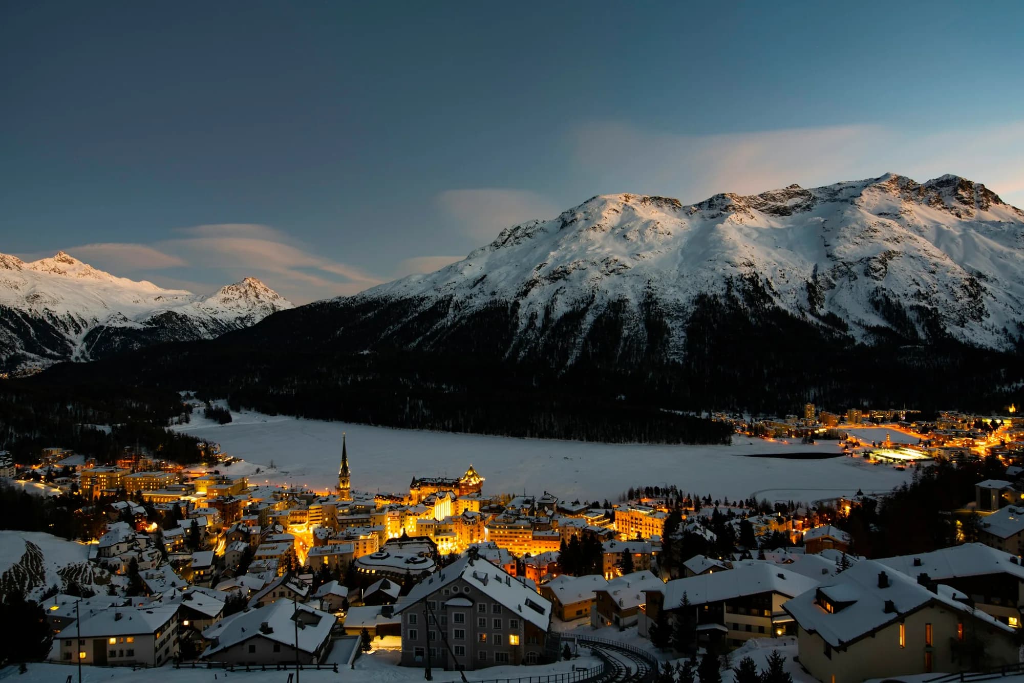 Snow-covered St. Moritz at dusk, with illuminated buildings beside the frozen lake and mountains behind.