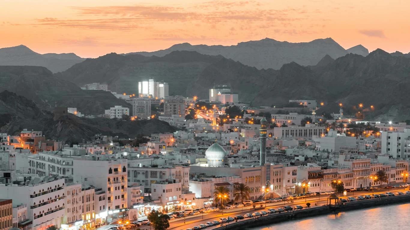 Muscat’s white, low-rise districts at dusk, between the Al Hajar mountains and the sea